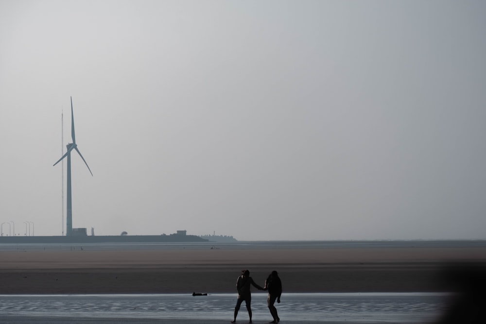 two people walking on a beach with a wind turbine in the background