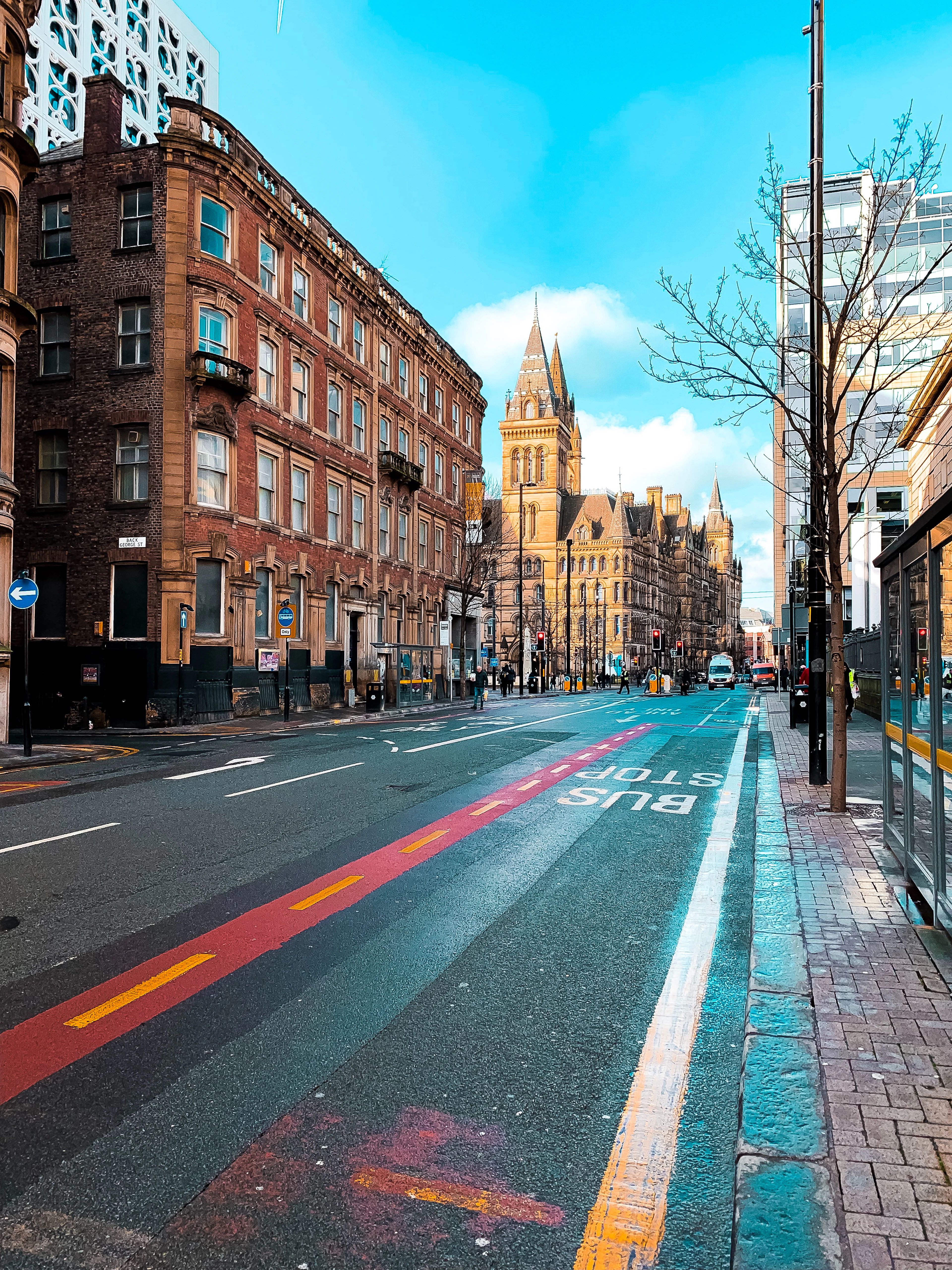 A wide central Manchester street lined with heavy Victorian red-brick and sandstone buildings, the gothic clock tower of Manchester Town Hall closing the view at the end under a bright blue sky.