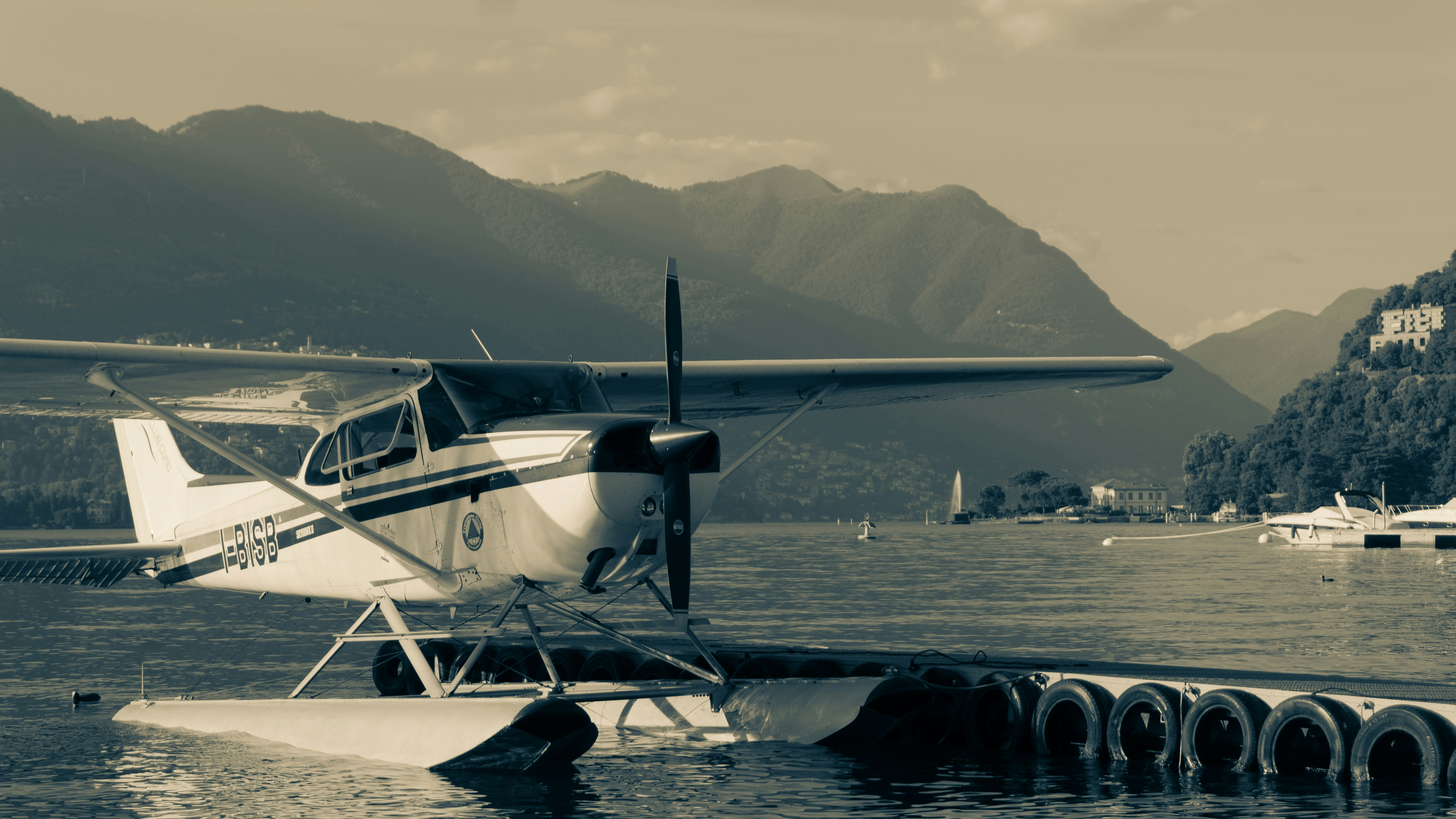 a small plane sitting on top of a body of water, Captured in timeless monochrome, this photograph exudes a nostalgic charm, featuring a seaplane serenely docked on the glassy waters of Lake Lugano. The sepia tones highlight the vintage elegance of the aircraft, juxtaposed against the tranquil backdrop of softly undulating mountains. This composition not only celebrates the fusion of technology and nature but also invites the viewer to ponder over the quieter, reflective moments of adventure and exploration. It
