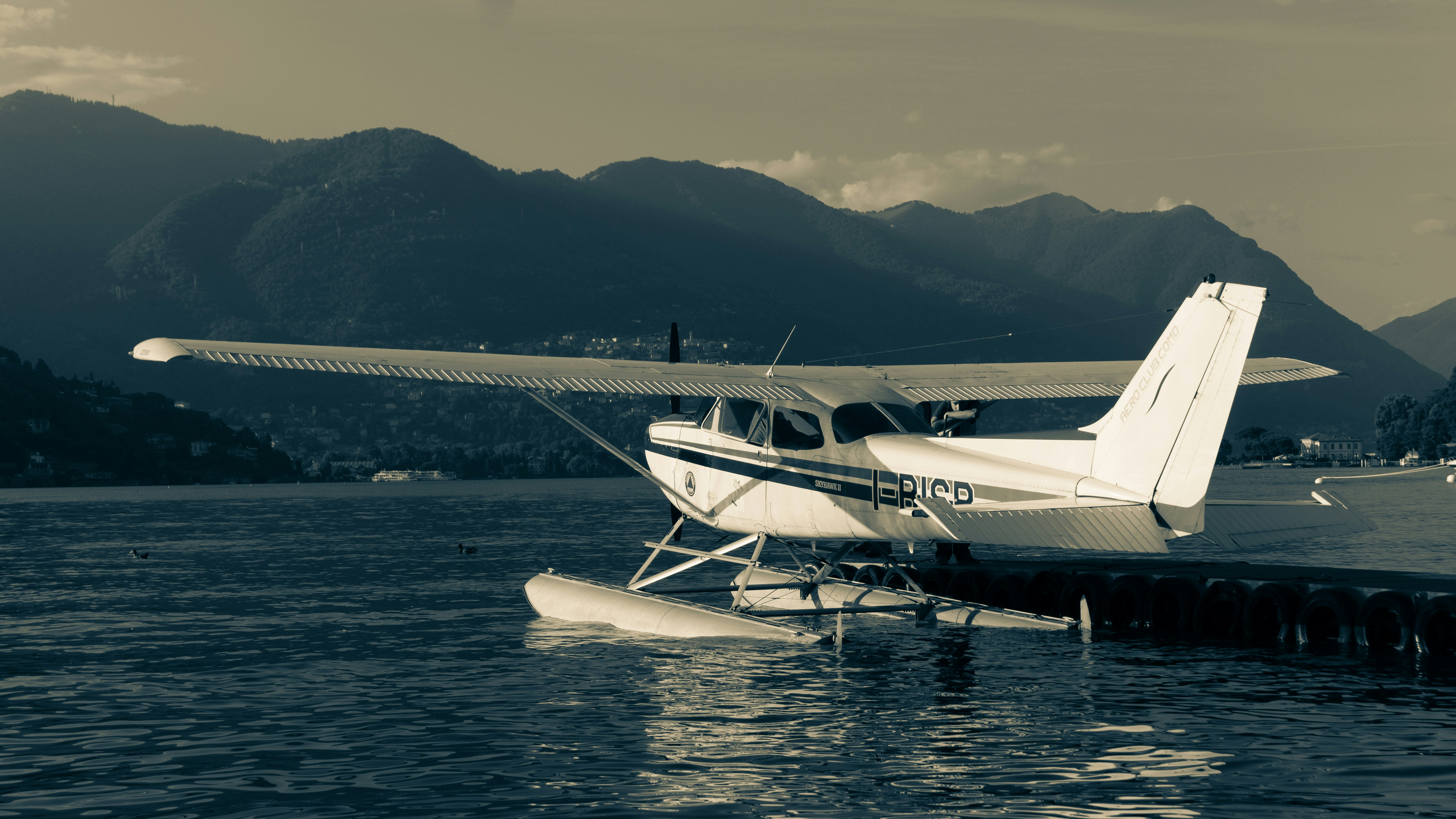 a small plane sitting on top of a body of water, In this monochrome image, a seaplane sits ready for departure on Lake Lugano, framed by towering mountains. A man resting against the aircraft brings a personal touch, embodying the calm before embarking on the next adventure. The photo melds the wanderlust of classic aviation with the raw beauty of the Swiss Alps, capturing a narrative of anticipation and the intimate quietude of travel.