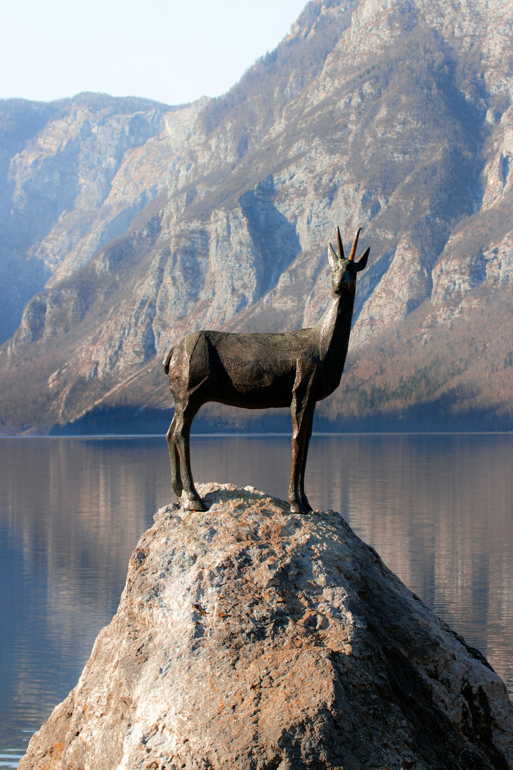 a statue of a goat stands on a rock in front of a mountain lake