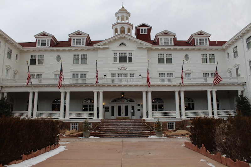 Front facade of a large white wooden hotel with red dormered roof, a central cupola, a columned veranda and a row of American flags, with patches of snow on the lawn