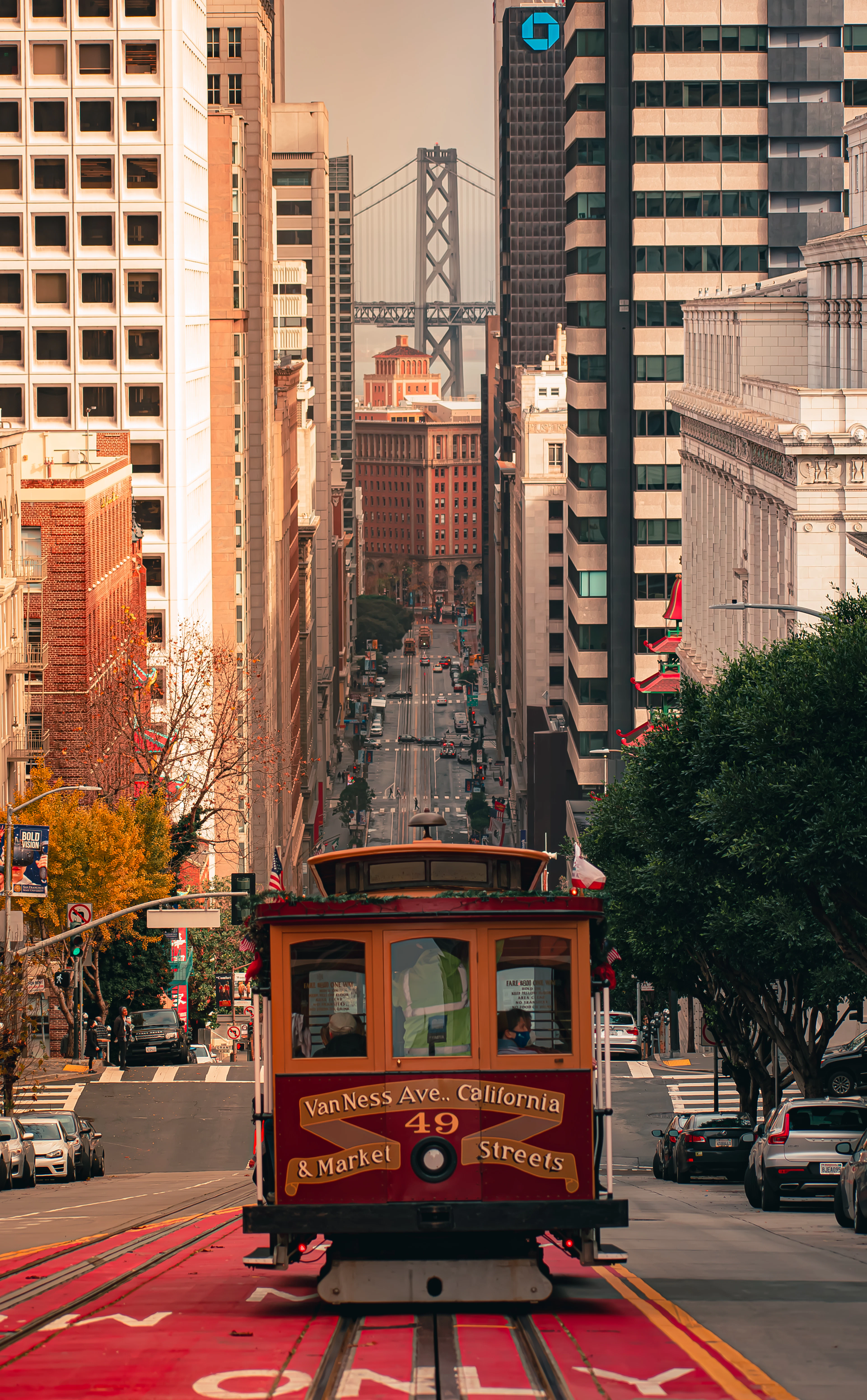 A San Francisco cable car lettered "Van Ness Ave, California & Market Streets" crests a steep hill, with the Bay Bridge rising between downtown towers behind it