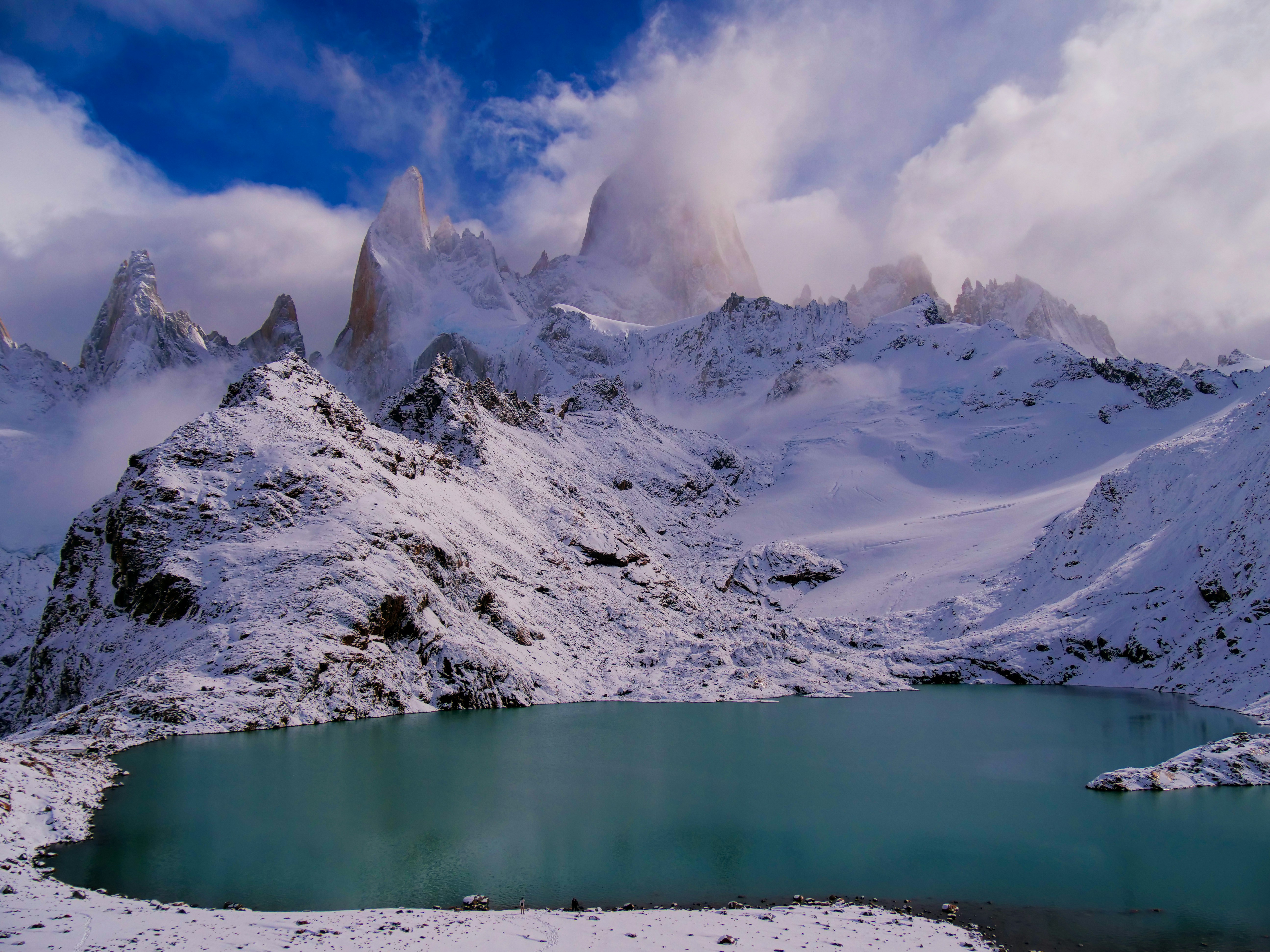 a lake surrounded by snow covered mountains under a cloudy sky
