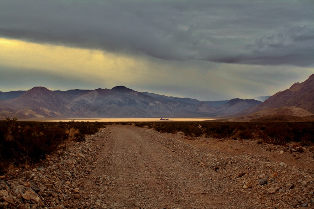 a dirt road surrounded by mountains under a cloudy sky