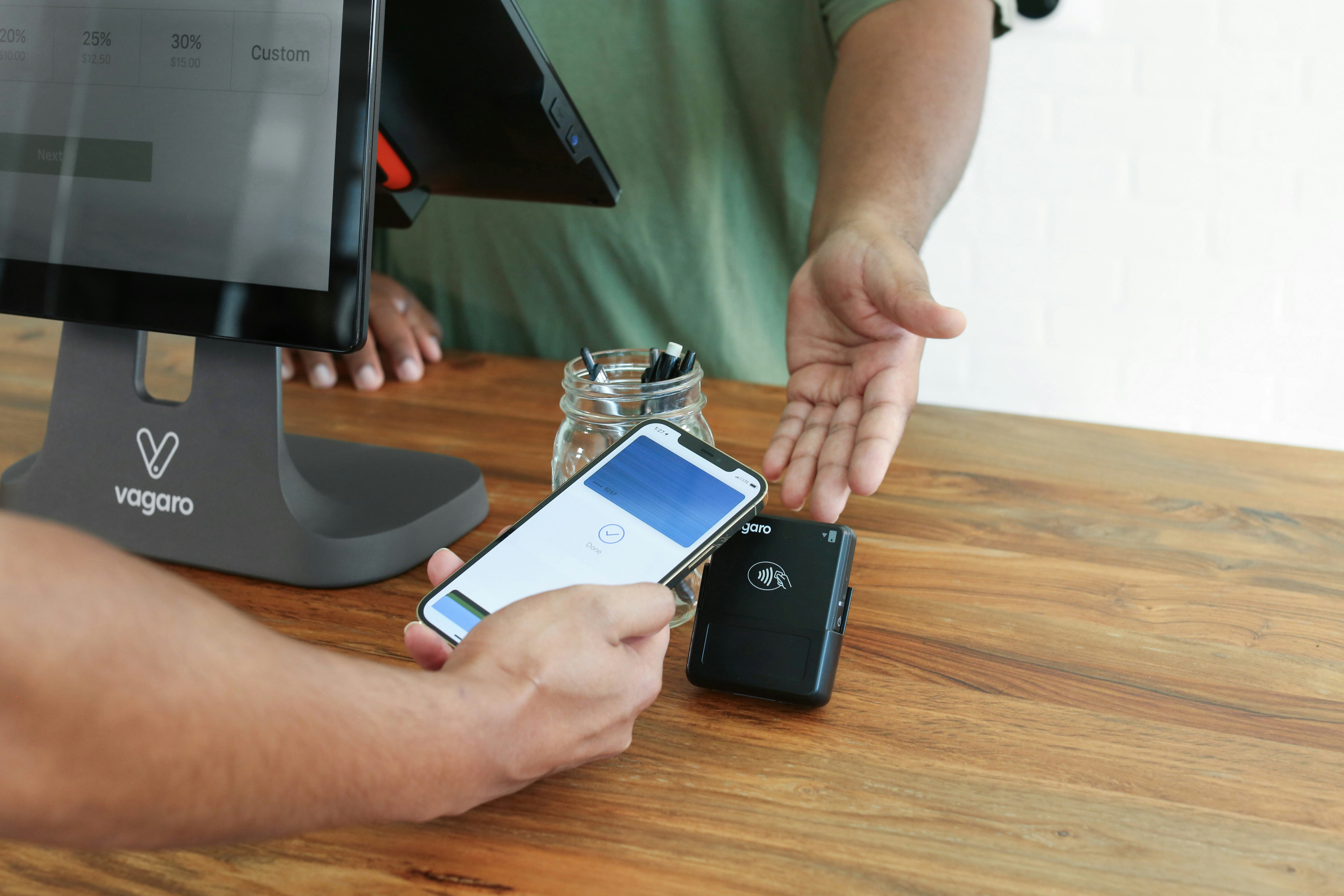 A person is holding an iPhone and using Apple Pay to checkout. The transaction is taking place with a Vagaro contactless card reader, positioned on a wooden table. Next to the card reader is a Vagaro point-of-sale (POS) system, creating a seamless and convenient payment setup.