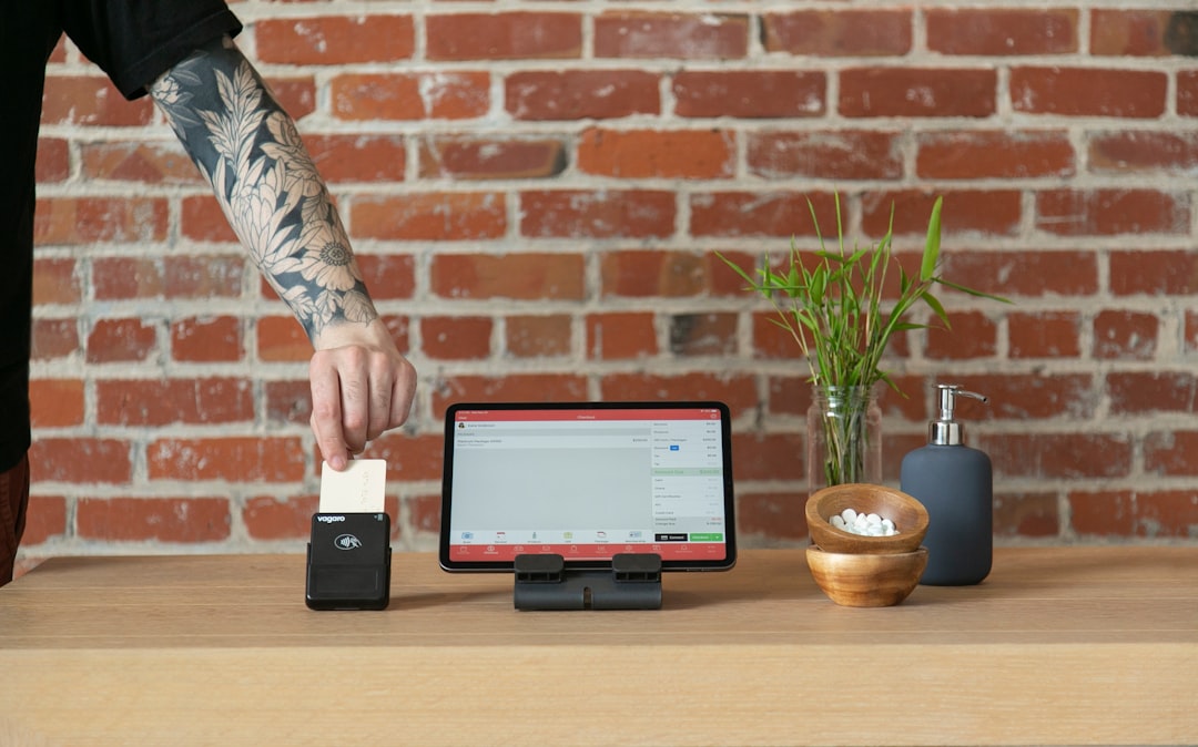 a man with a tattooed arm standing over a computer, A person is making a transaction with a credit card on a Vagaro contactless card reader. The sleek and modern payment process introduces a touch of convenience, enhancing the overall checkout experience.