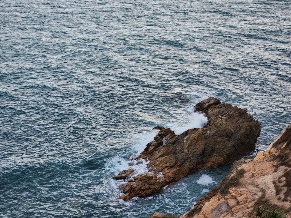 The image depicts a rocky cliff overlooking the ocean, with the vast expanse of water stretching out to meet the sky at the horizon. The deep blue-green color of the water contrasts with the brown hues of the rocks and cliffs. A few small waves are crashing against the rocks on the right side of the image, adding a dynamic element to the scene.