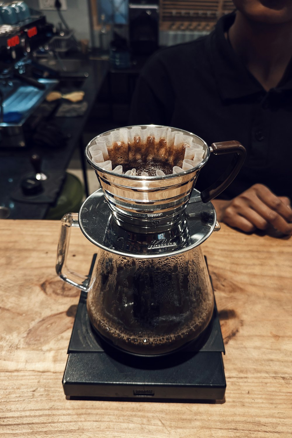 a coffee pot on a wooden table with a person in the background