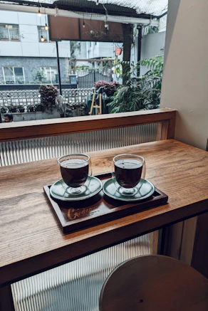 two cups of coffee sit on a tray in front of a window
