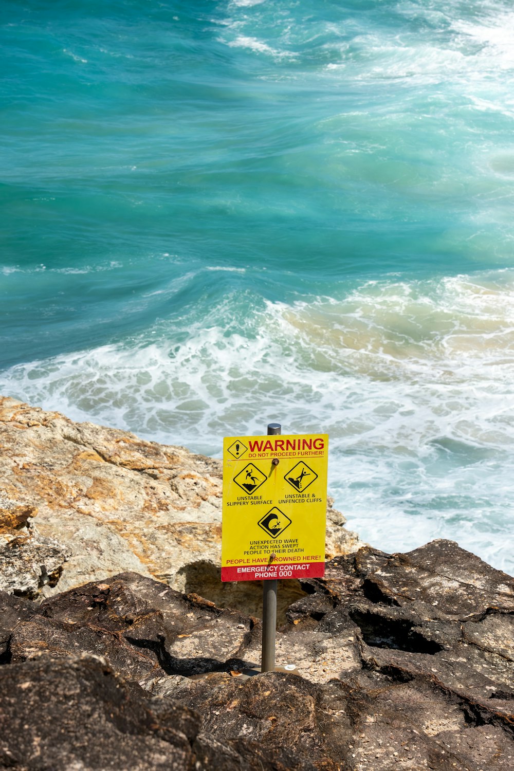 a yellow warning sign sitting on top of a rock near the ocean