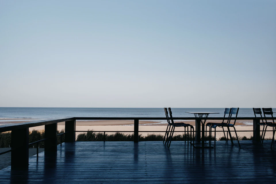The image depicts a serene beachfront patio with a wooden deck and metal railing, overlooking the vast ocean. The deck is adorned with six black chairs arranged around a round table, facing the water. The table is positioned on the right side of the deck, while four additional chairs are placed along the left side. The chairs appear to be empty at the moment but can be occupied by guests enjoying the view or engaging in conversation.