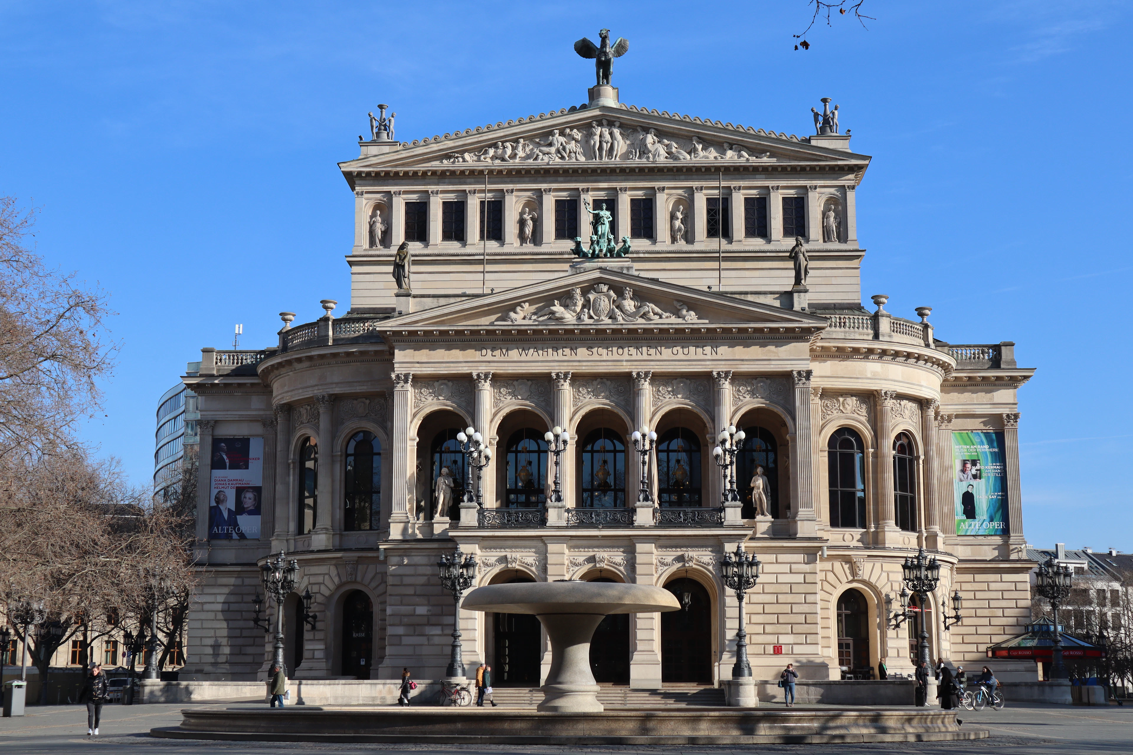 The pale sandstone facade of Frankfurt's Alte Oper, its statue-lined pediment and arcaded loggia crisp against a deep blue winter sky, with the low stone bowl of the Lucae fountain standing on the near-empty Opernplatz in front.