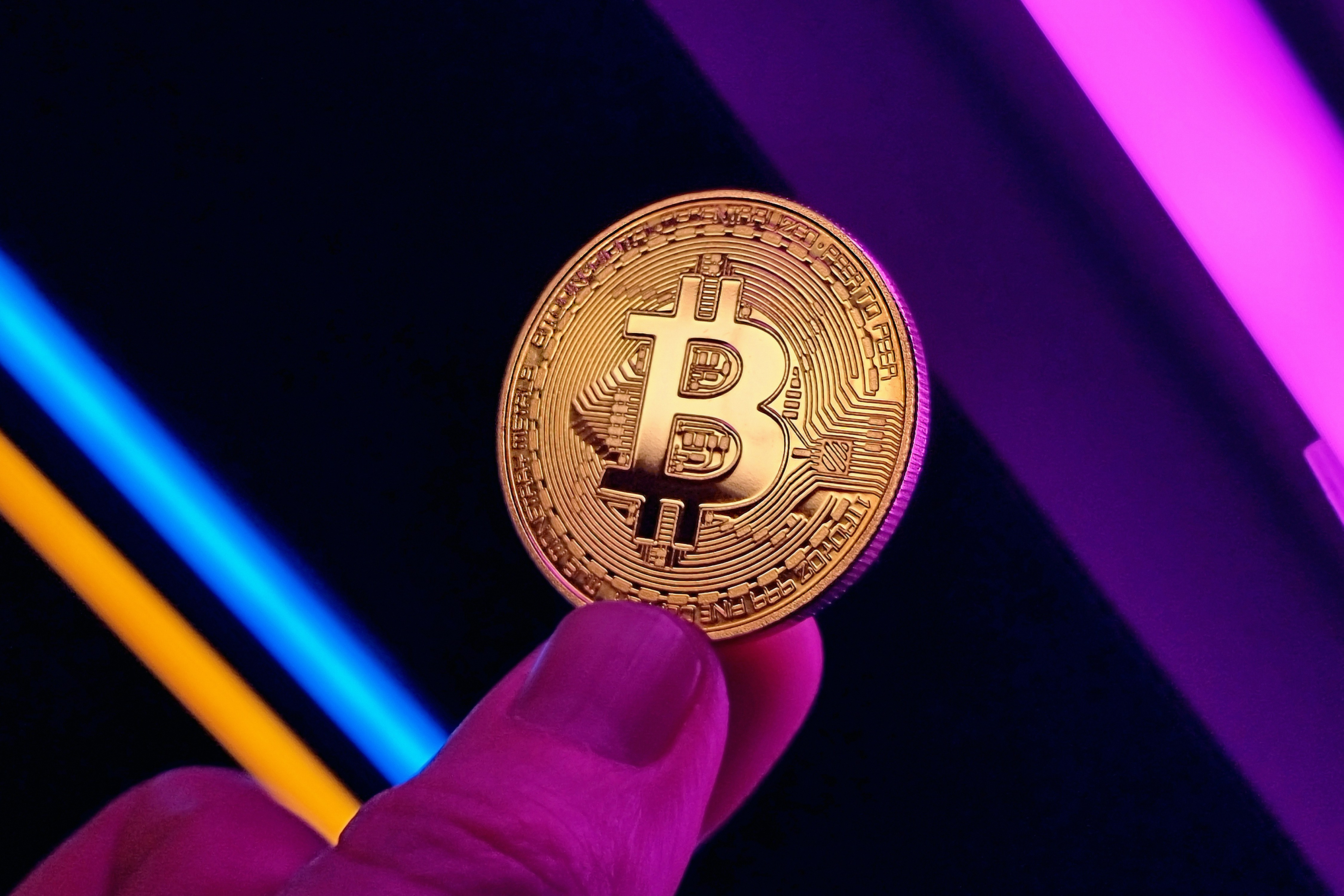 Young man holding gold Bitcoin cryptocurrency coin between his fingers, against ambient neon lights. Purple, blue and orange lights in the background.