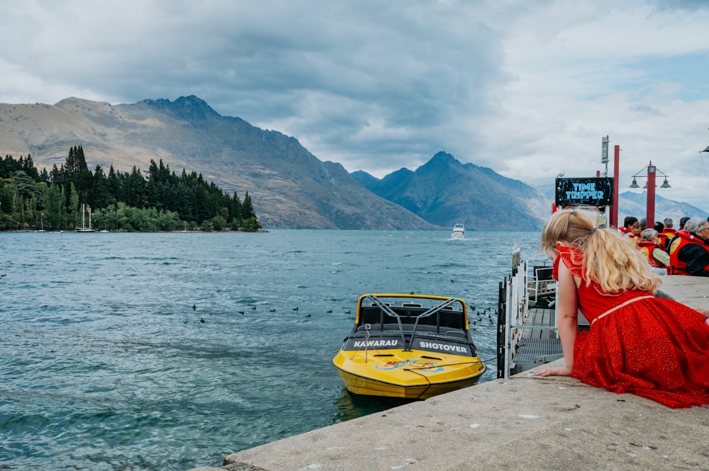 una mujer con un vestido rojo sentada en un muelle junto a un barco amarillo