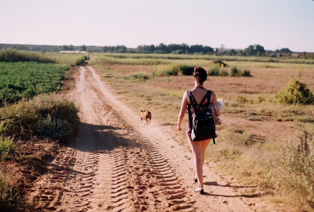a woman walking down a dirt road next to a dog