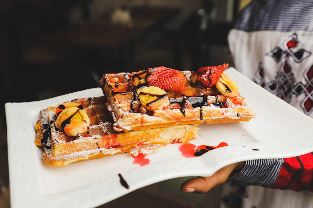 a person holding a plate of waffles with fruit on top