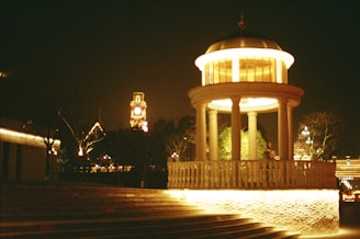 a gazebo lit up at night with a clock tower in the background