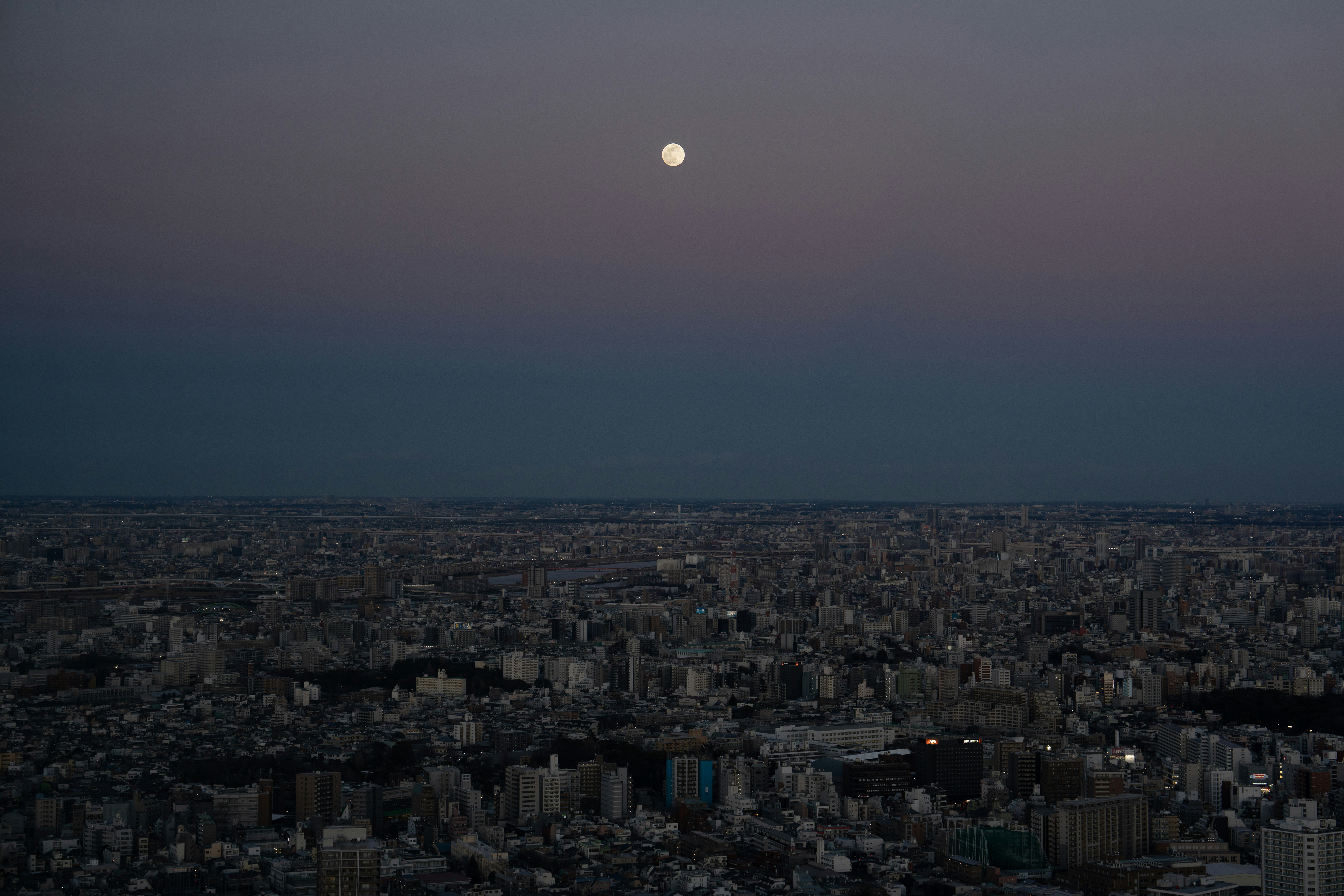 Dusk cityscape photograph with a full moon rising above a densely packed urban skyline, lights beginning to glow as darkness settles.