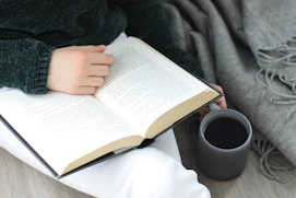 a person holding a book and a cup of tea with cozy blanket