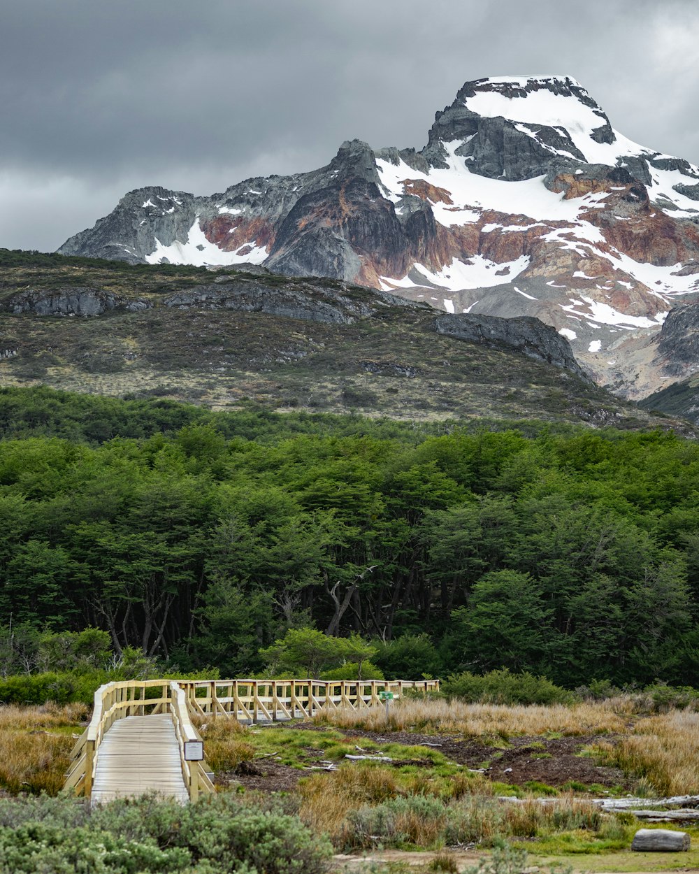 a wooden bridge crossing a stream in front of a snow covered mountain