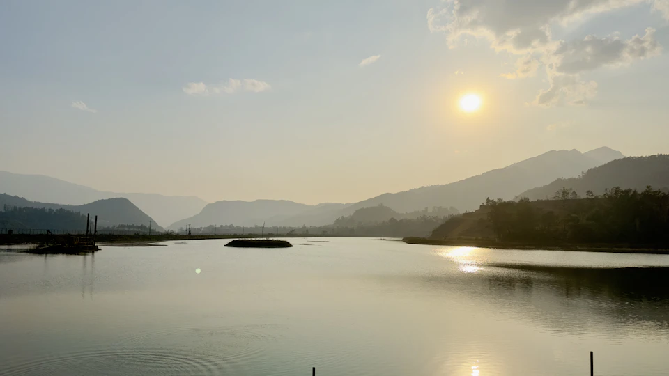 The image depicts a serene lake surrounded by mountains, with the sun setting behind them and casting a warm glow over the scene. The sky is painted in shades of blue, dotted with fluffy white clouds that add depth to the landscape. The water of the lake reflects the vibrant colors of the sunset, creating a beautiful mirror-like effect. In the foreground, there are two small islands visible on the left side of the image.