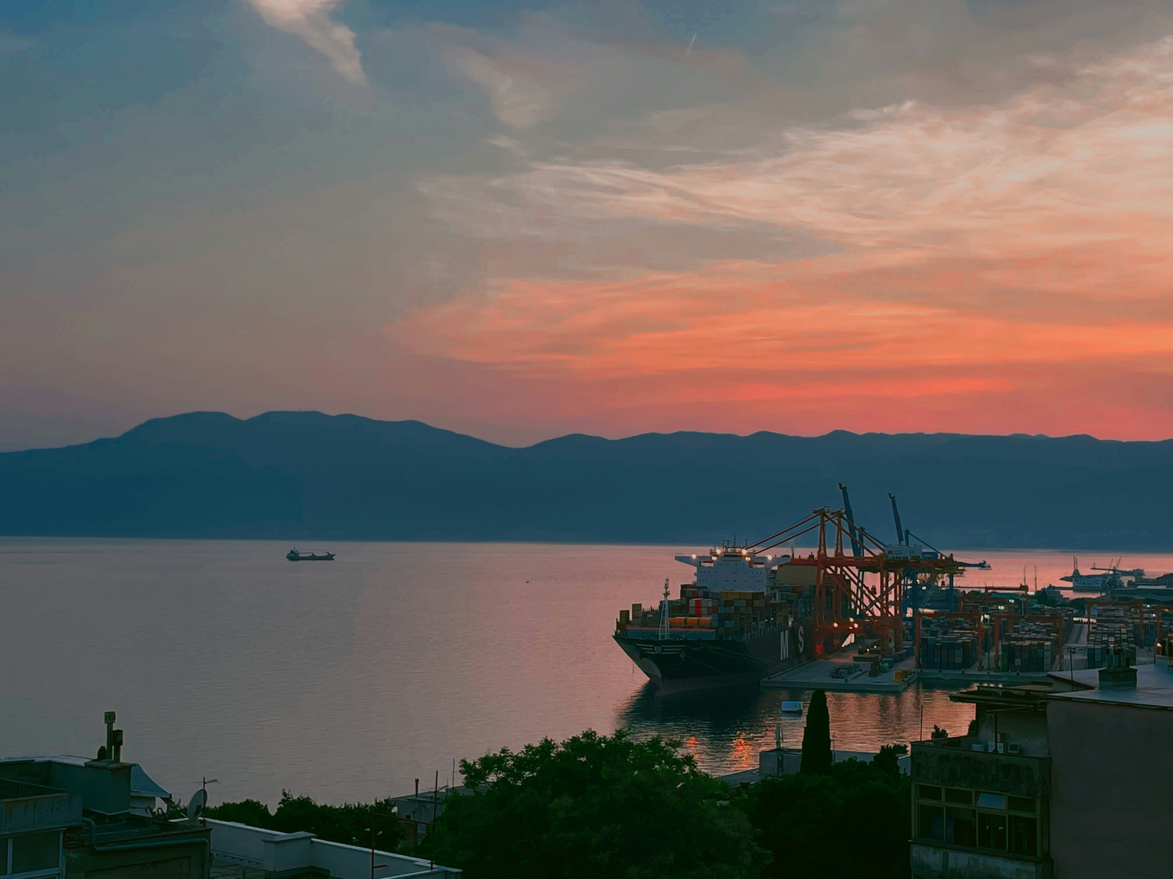 Container ship loaded with cargo containers at sunset in a busy port, calm dark water reflecting orange sky