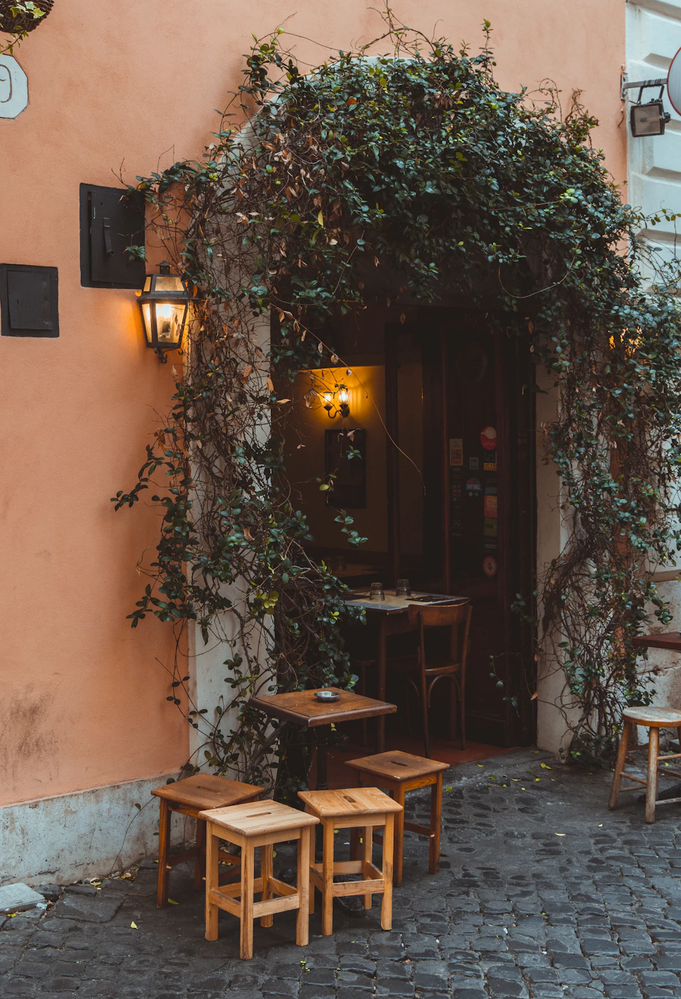 The image depicts a quaint outdoor café with a warm, inviting atmosphere. The focal point is an archway adorned with lush green vines and plants, leading to a wooden door that opens up to a small patio area furnished with two tables and four chairs. Each table is accompanied by a matching chair, creating a harmonious balance in the scene. The tables are positioned on either side of the archway, while the chairs are arranged around it. A lamp post stands tall at the entrance, providing ample lighting for the outdoor seating area.
