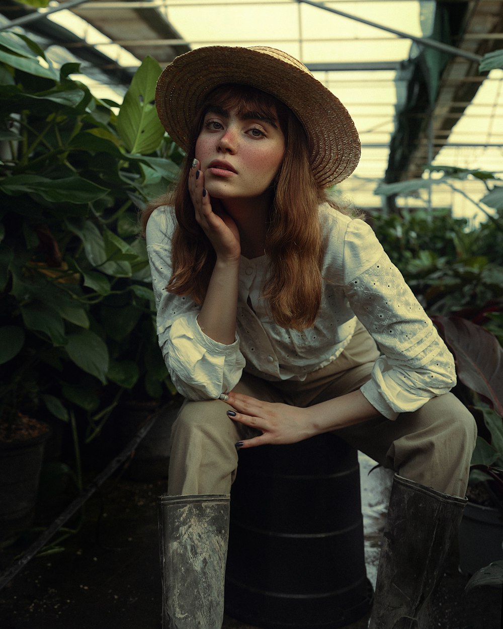 a woman in a hat sitting on a bucket in a greenhouse