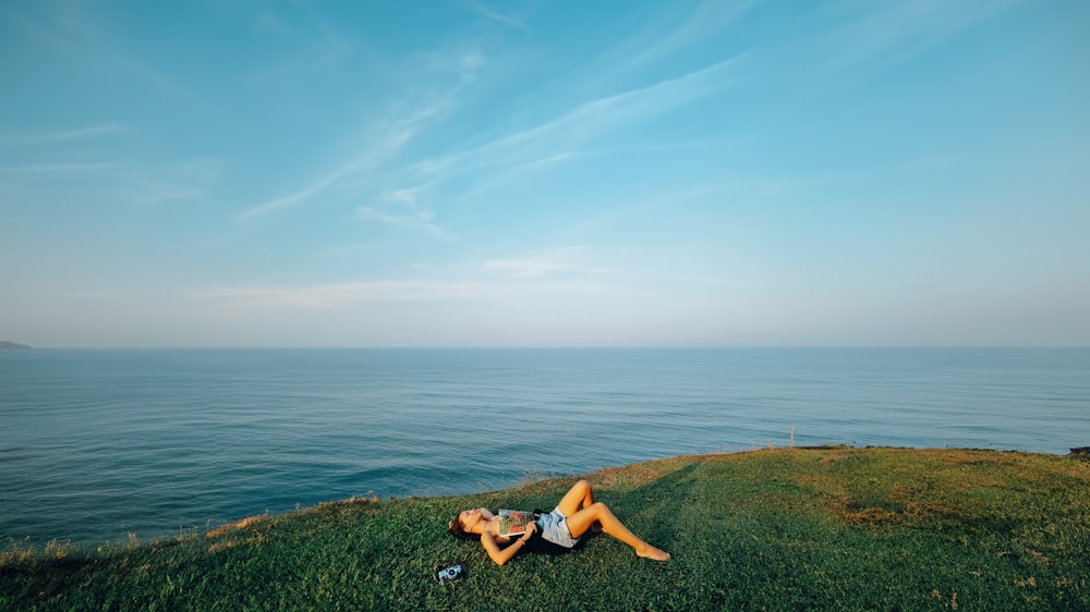 a woman laying on top of a lush green hillside next to the ocean