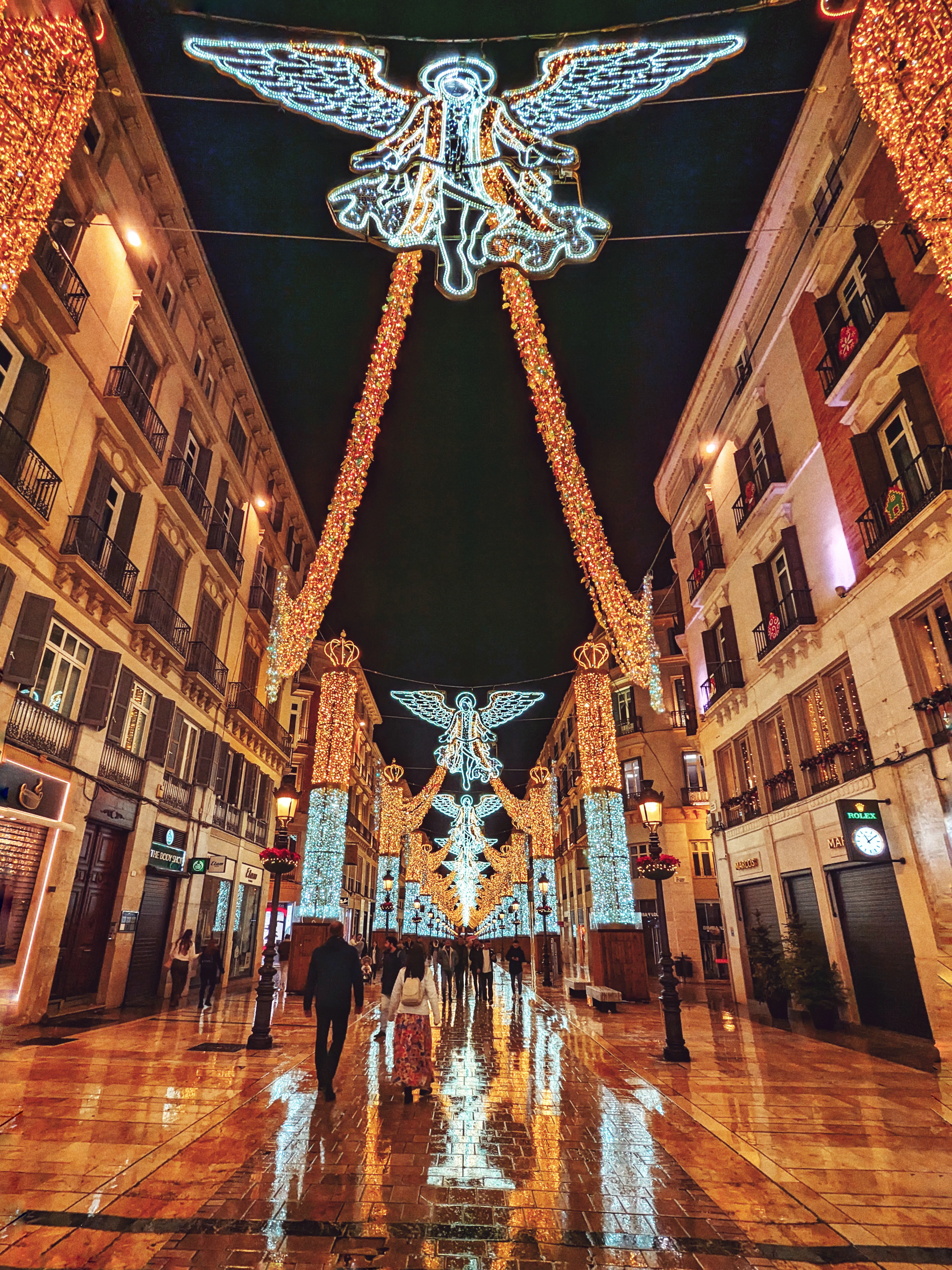 Calle Larios in Malaga at Christmas, giant illuminated angels arching over the street between the balconied buildings and their light reflected in the wet marble paving as people walk beneath