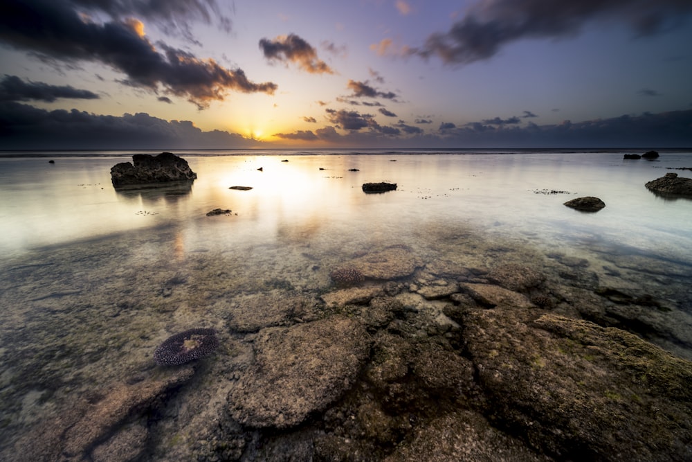 the sun is setting over the ocean with rocks in the water