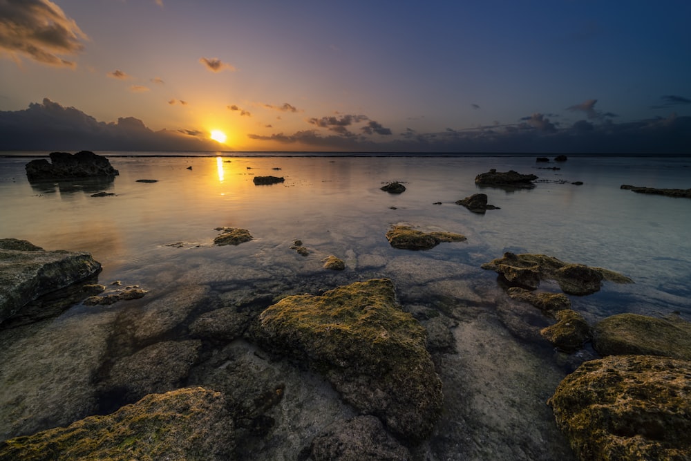 the sun is setting over the ocean with rocks in the foreground