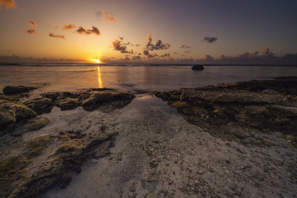 the sun is setting over the ocean with rocks in the foreground
