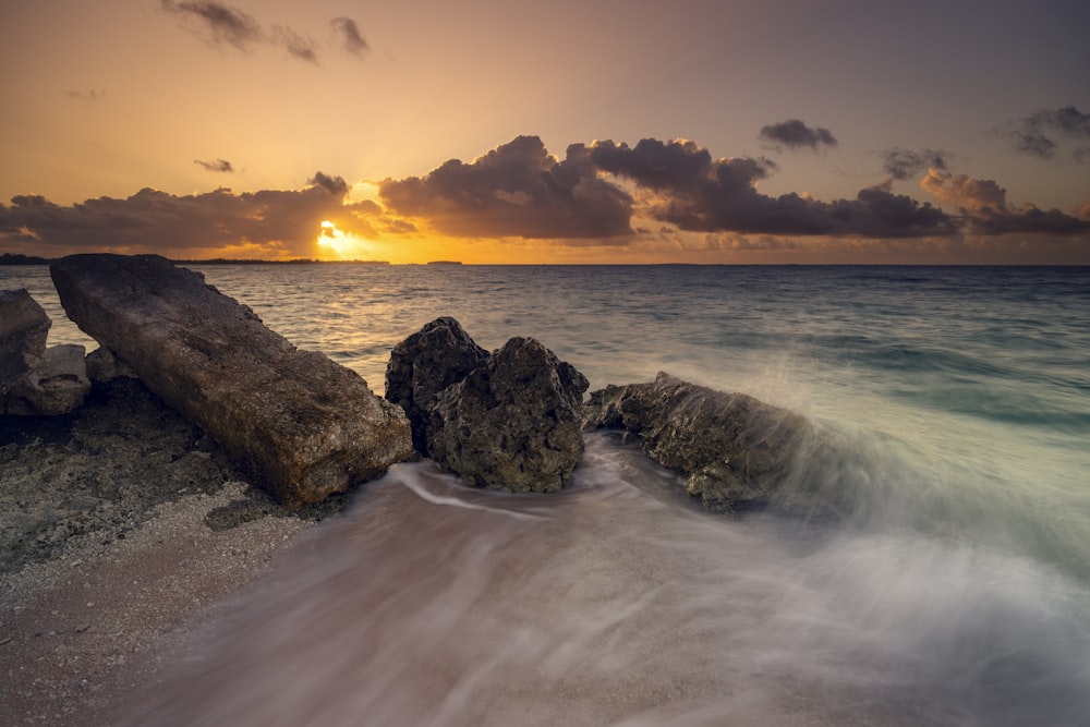 the sun is setting over the ocean with rocks in the foreground