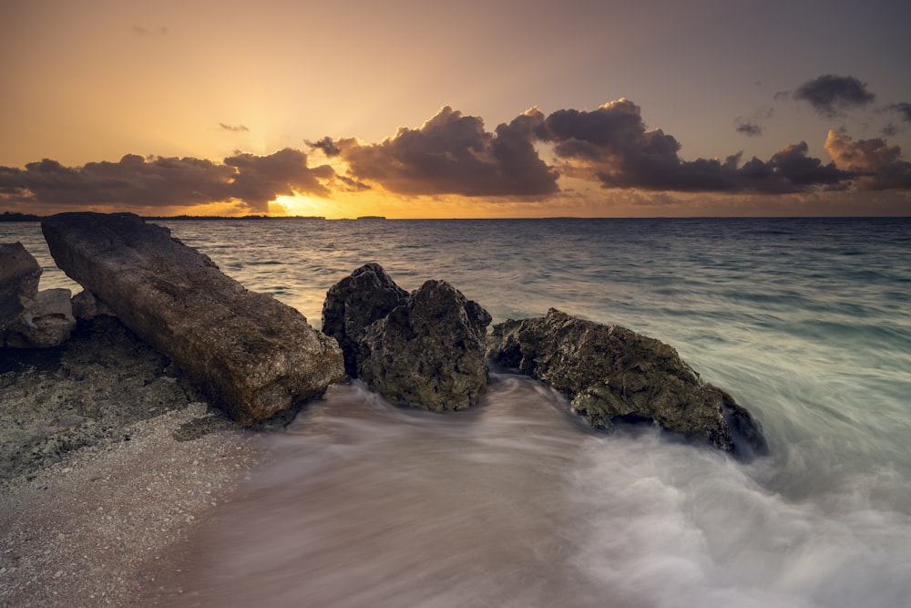 the sun is setting over the ocean with rocks in the foreground