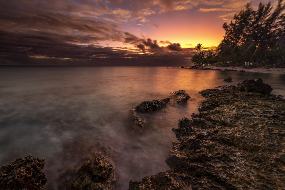 the sun is setting over the ocean with rocks in the foreground