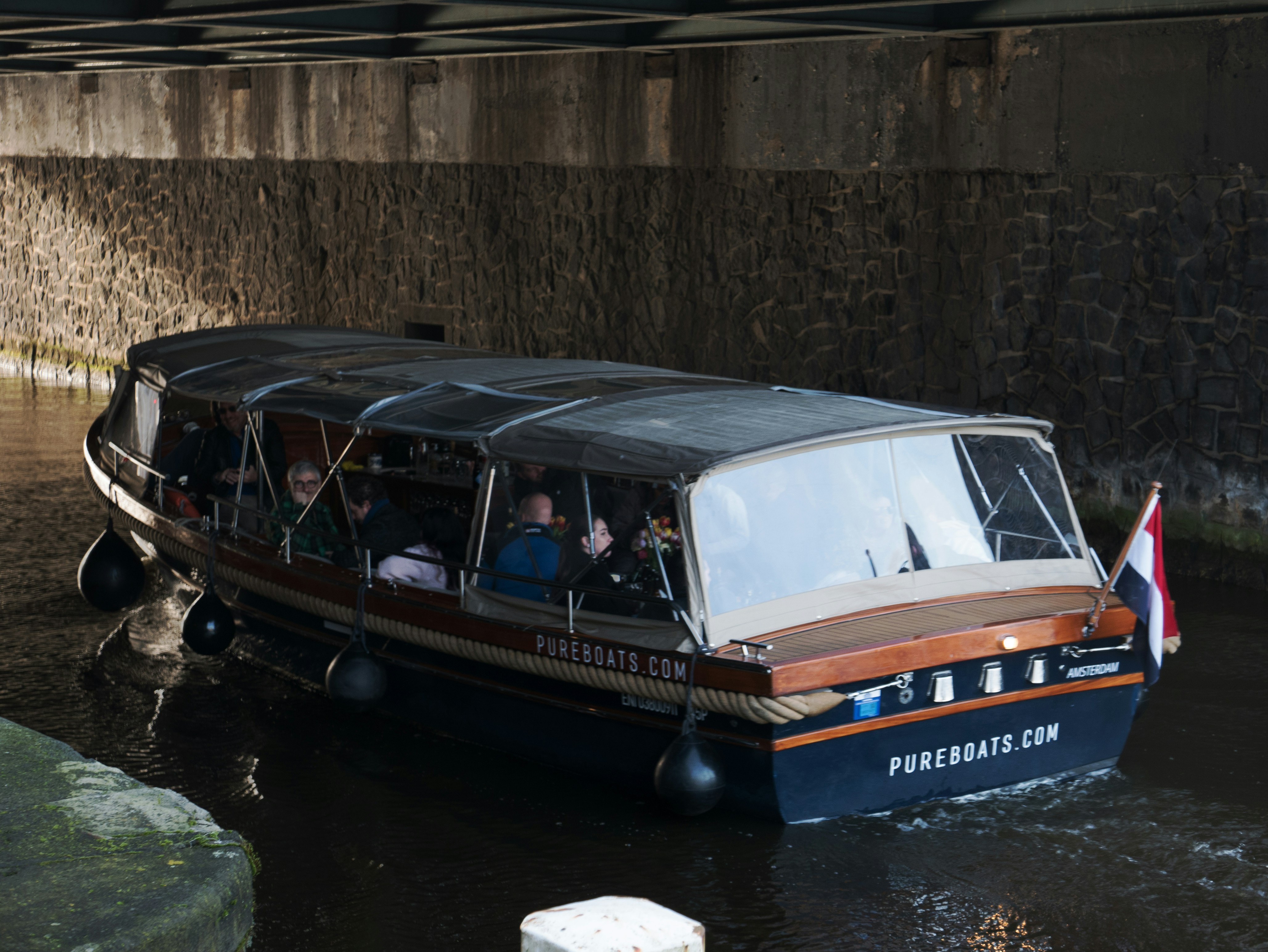 a boat with passengers going under a bridge, A tourist boat is under the bridge of the road Prins Hendrikkakde in Amsterdam city. Amsterdam is a canal city with a lot of tours and trips through the whole old city center. I like the play here of the light and shadows and reflections of the sunlight in the glass windows. Street photography of canal boats in Amsterdam city by Fons Heijnsbroek; free download urban photo, The Netherlands, 2024