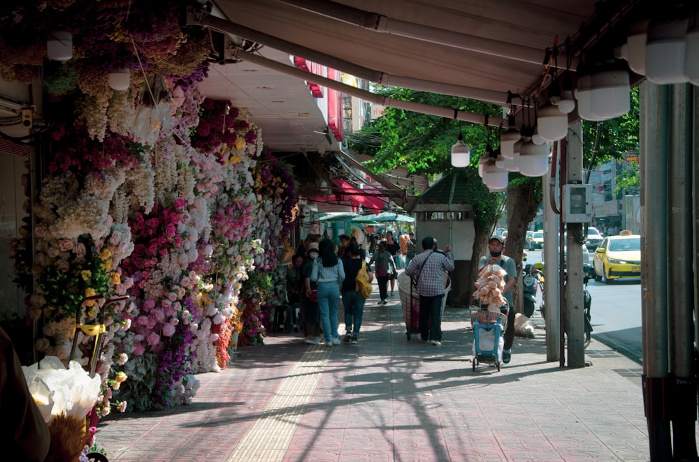 a group of people walking down a street next to a wall of flowers