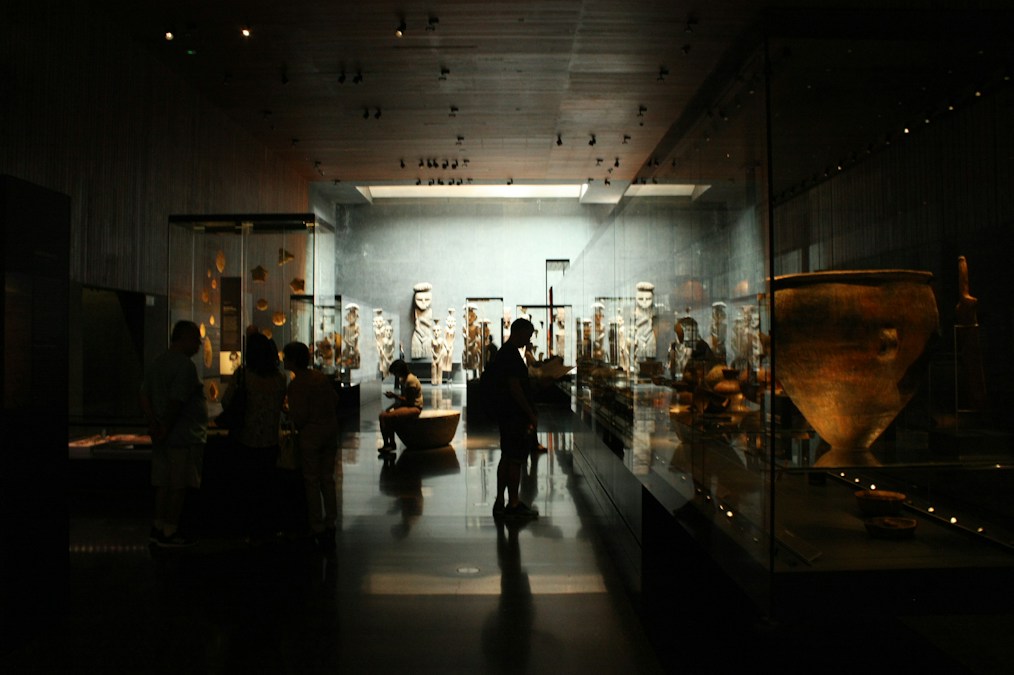 Des visiteurs en silhouette déambulent le soir dans une salle de musée tamisée, entre statues et vitrines éclairées