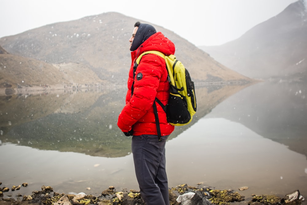 a man in a red jacket and yellow backpack looking at a lake