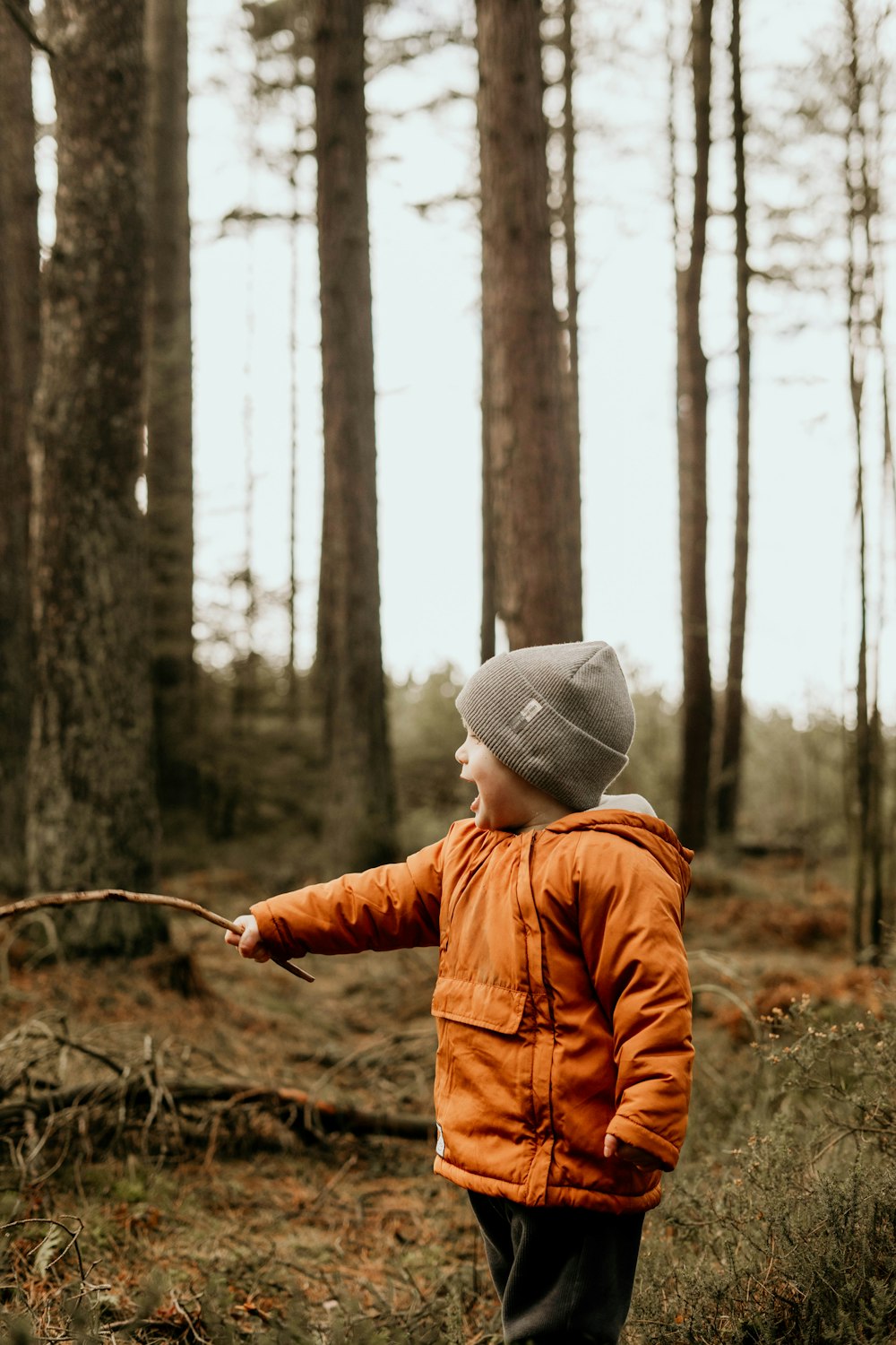 a young boy in an orange jacket is holding a stick in the woods