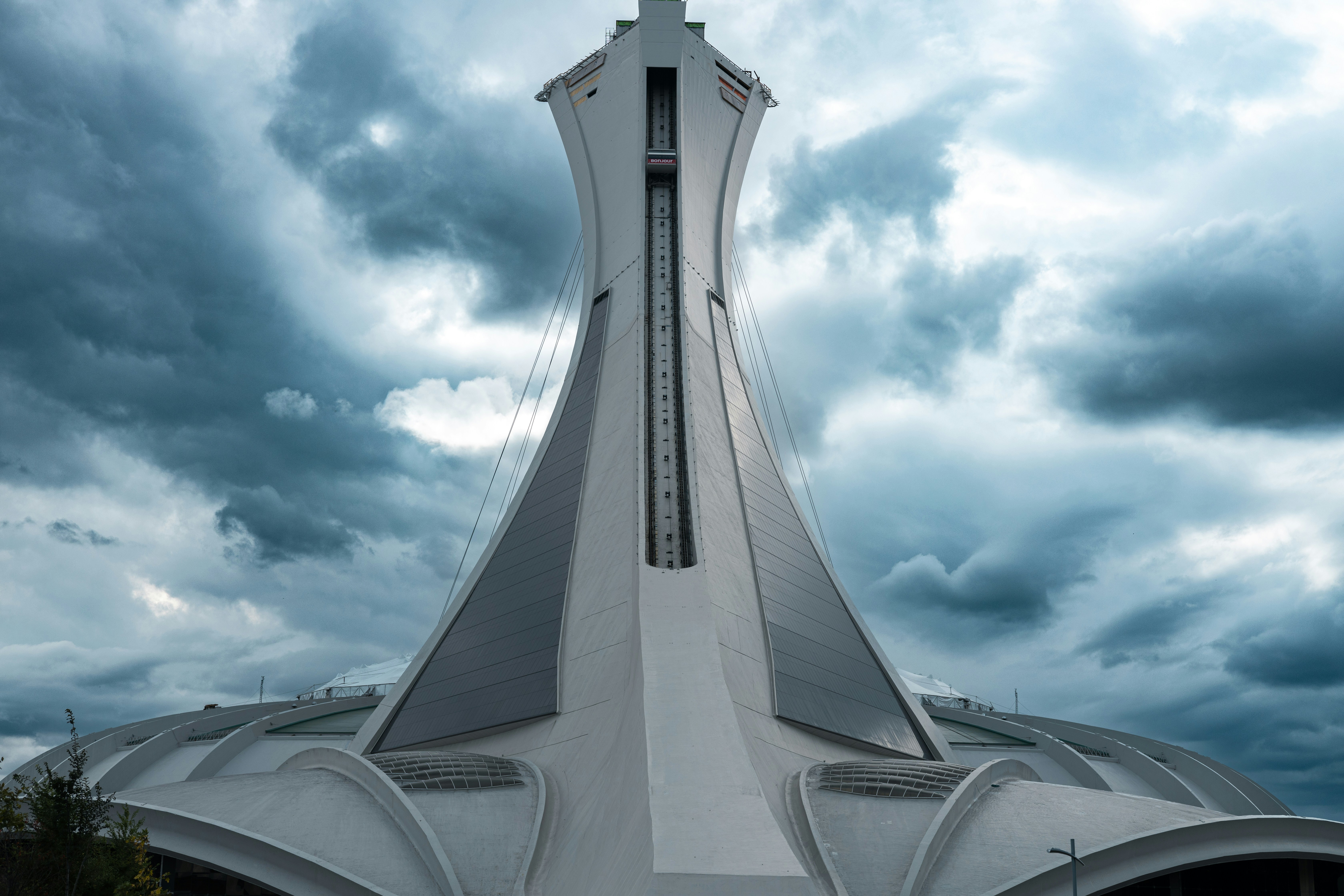 This striking photograph captures the iconic Olympic Stadium in Montreal, standing tall against a backdrop of moody, stormy skies. The stadium's unique, inclined tower, the world's tallest at 175 meters, dramatically stretches towards the clouds, emphasizing the bold architecture that symbolizes Montreal's innovative spirit. The contrasting light and dark elements in the image enhance the futuristic silhouette of the structure, while the foreboding sky adds a sense of impending majesty and power.