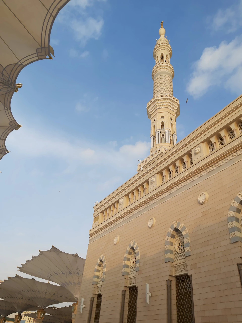 The image depicts a large, light-colored stone mosque with a tall minaret and multiple arched windows on its facade. The mosque is situated under a clear blue sky dotted with white clouds. In front of the mosque, there are several white umbrellas providing shade to visitors or passersby.