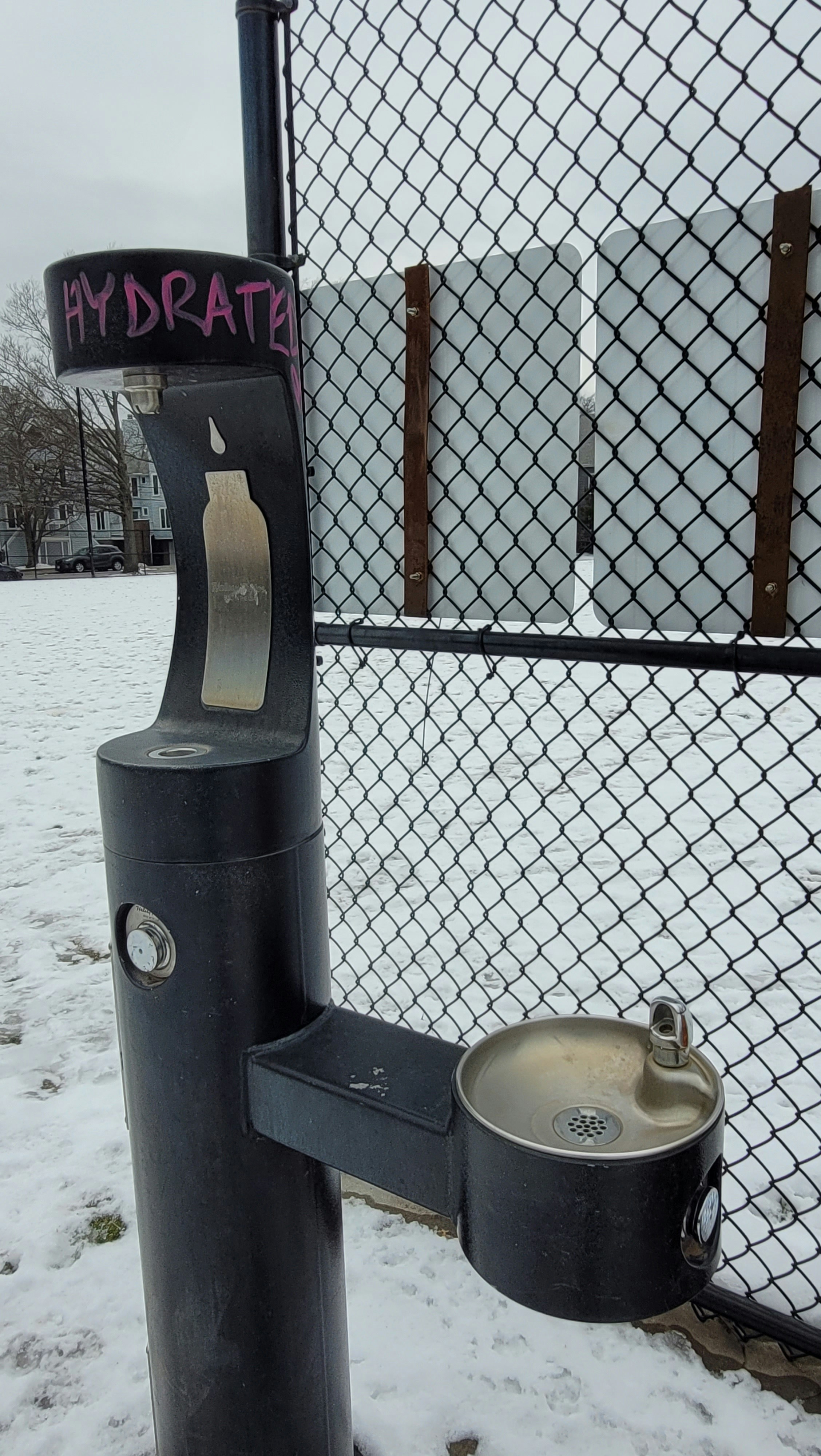 A water fountain stands in a snowy park. Graffiti on it reads, \