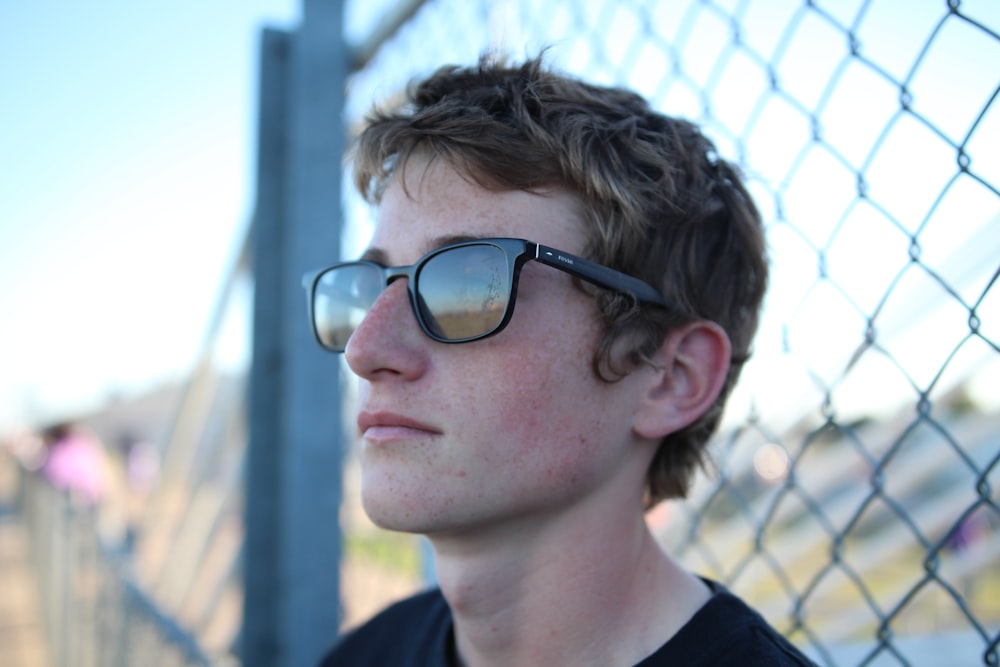 a young man wearing sunglasses standing next to a fence