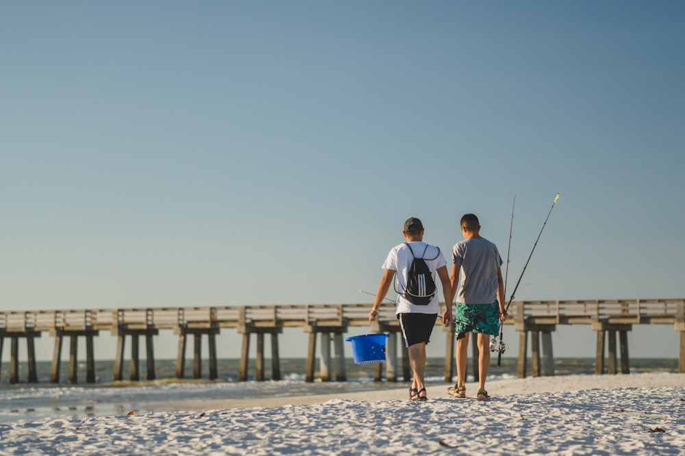 a couple of men walking down a beach next to a pier