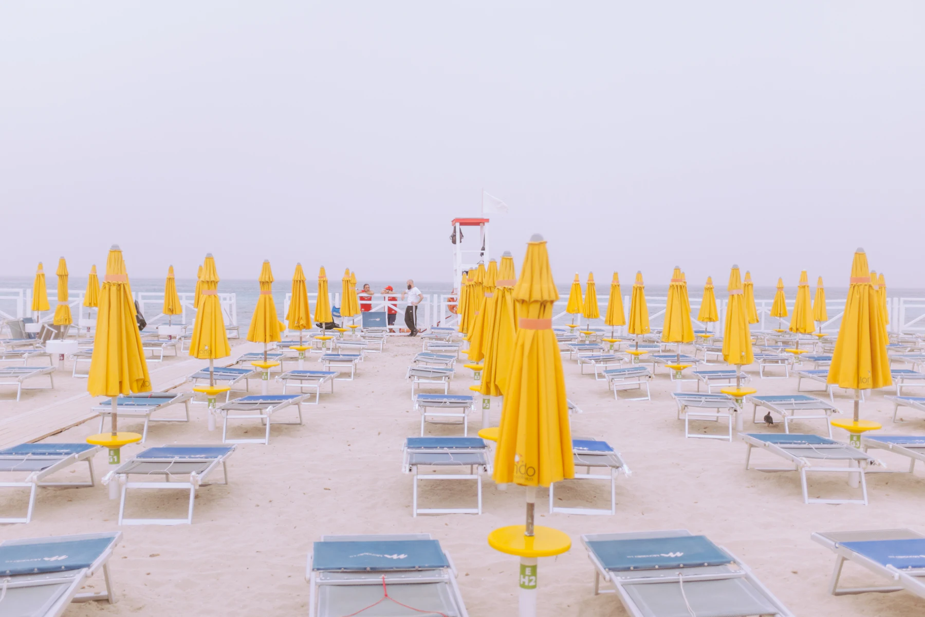 Closed yellow parasols and blue loungers lined up on a pale beach, lifeguard post