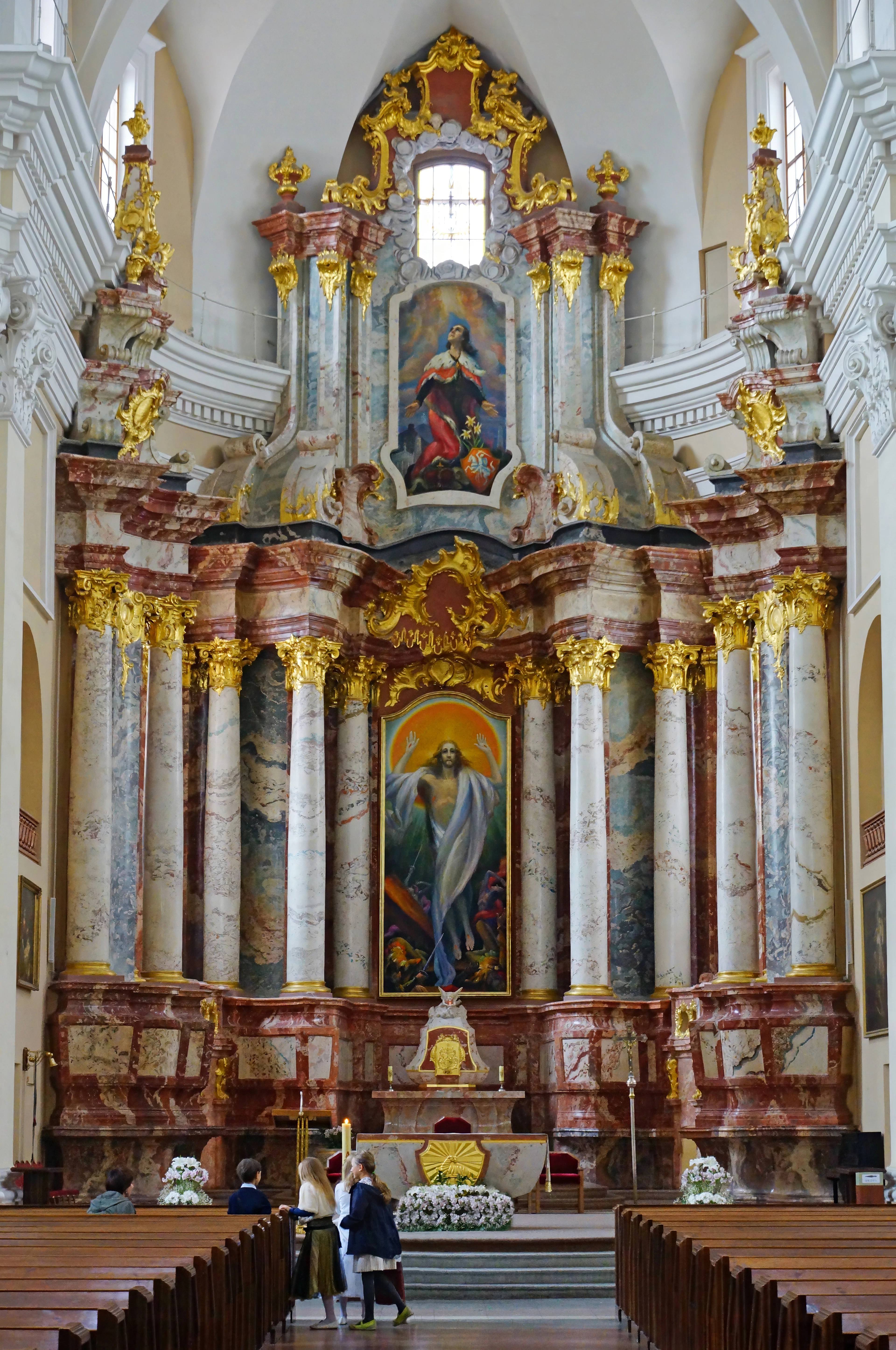 The marble and gilt high altar of a baroque church in Vilnius rising above empty pews, columns of veined red and grey stone flanking a painting of the risen Christ, with a few visitors standing at the altar rail.