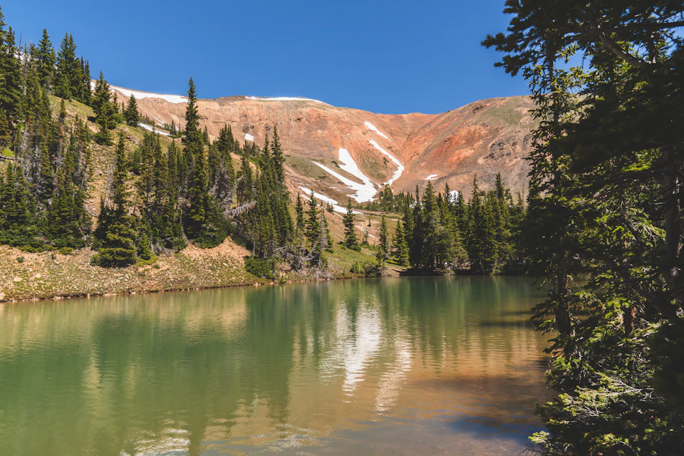 The image depicts a serene mountain lake nestled amidst lush greenery. The lake, with its vibrant green color and calm waters, is surrounded by an array of trees and shrubs that add to the natural beauty of the scene. In the distance, a majestic mountain range can be seen, providing a stunning backdrop for this picturesque landscape.

The sky above is a clear blue, suggesting a bright and sunny day. The image is taken from a low angle, which enhances the grandeur of both the lake and the mountains in the background.